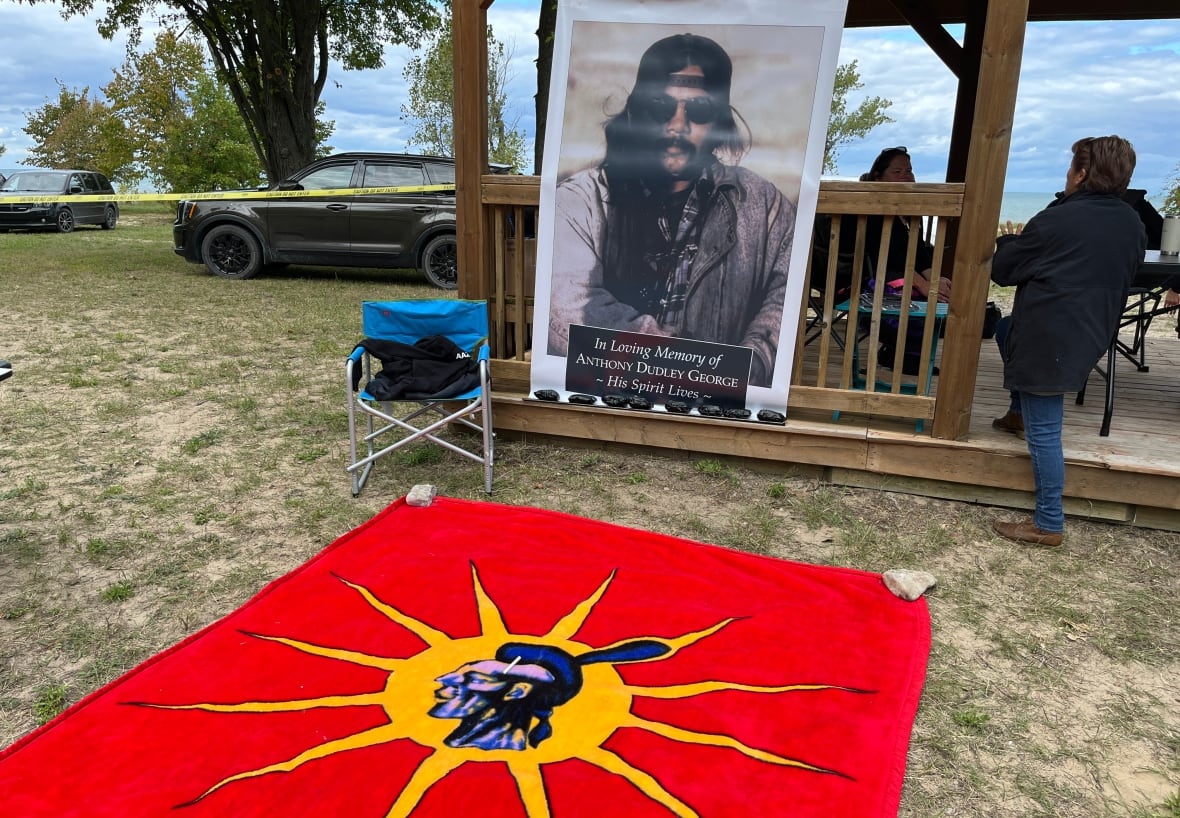 A picture of Dudley George stands behind a blanket with the red and yellow warrior flag on it.