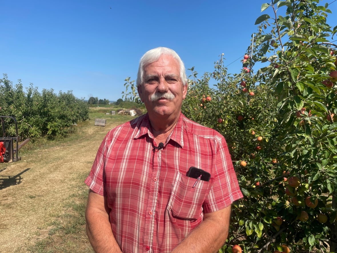A man with white hair and a white mustache poses in front of an apple orchard.