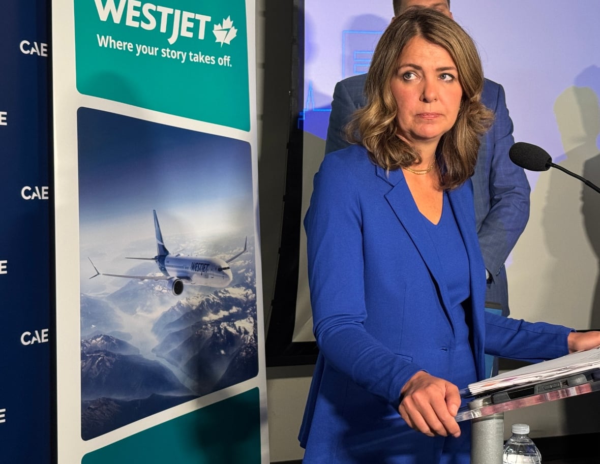Danielle Smith stands in front of a podium. She is a white woman with light brown hair that falls around her collarbone. She is wearing a monochromatic royal blue suit and looking to her right. She is standing in front of a WestJet poster.