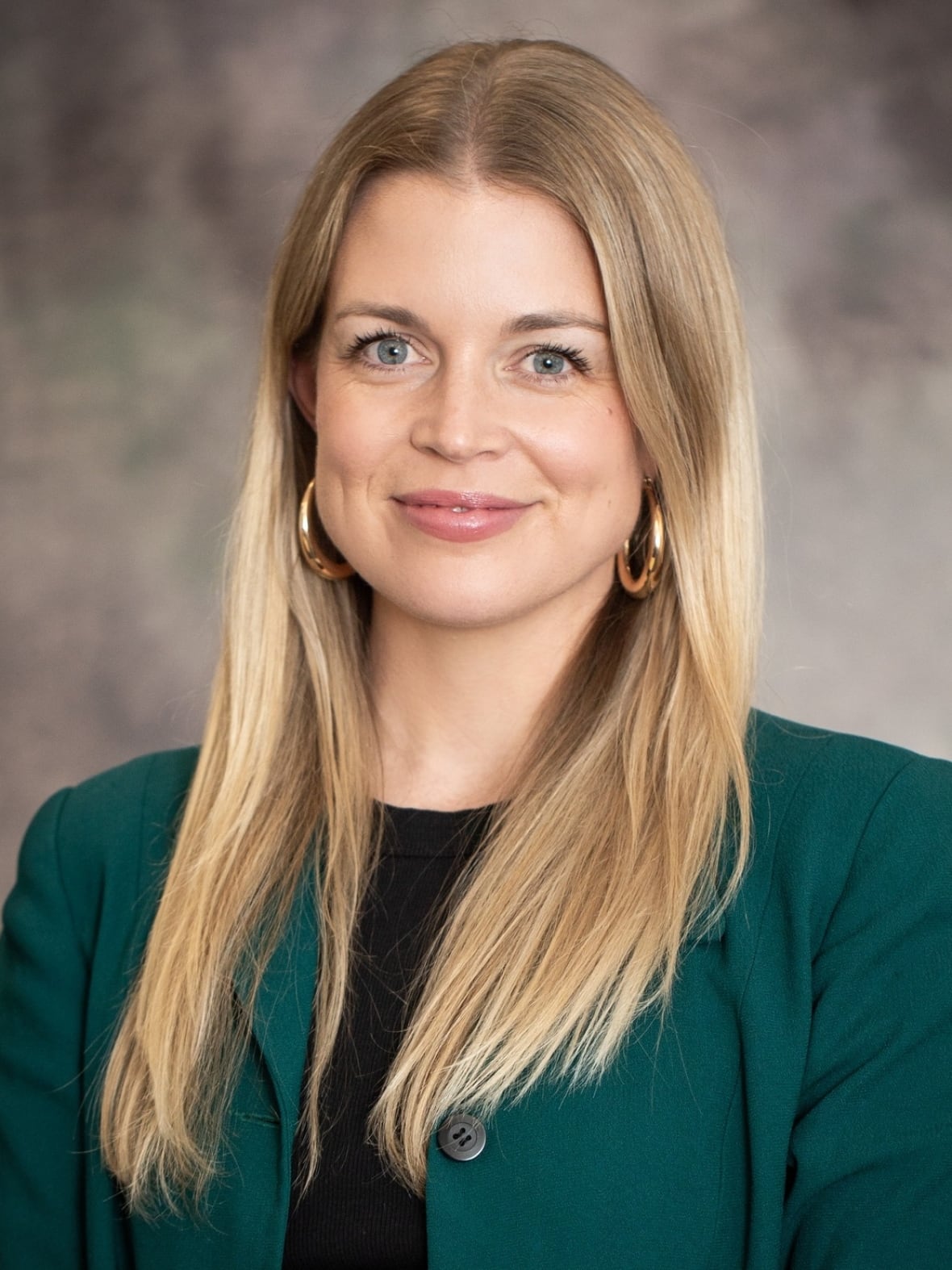 A woman with blonde hair poses for a portrait. She wears a green blazer and black shirt. 