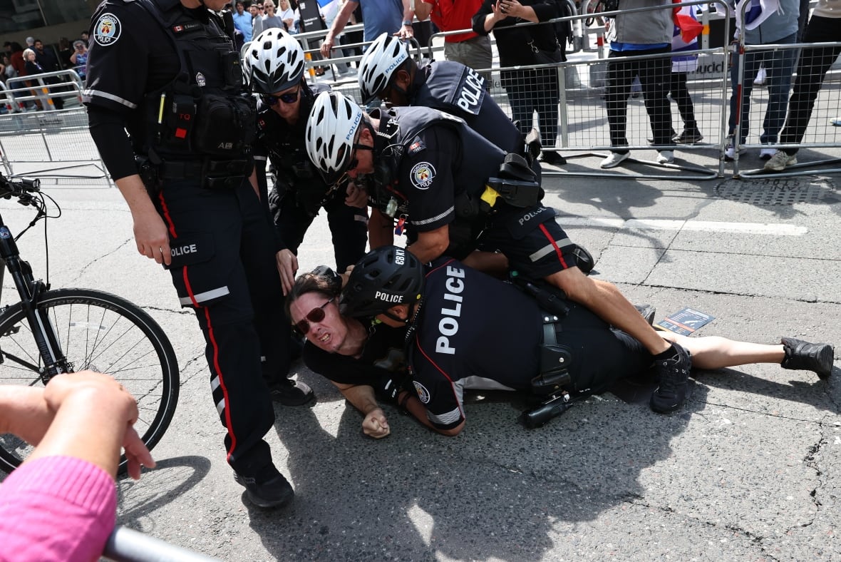a man on the ground wearing sunglasses lays struggling under four police officers who pin him down. a fifth officer stands on the left side. in the background protesters can be seen standing behind a barricade