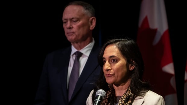 A woman speaks into a microphone as a man in a suit stands beside her.