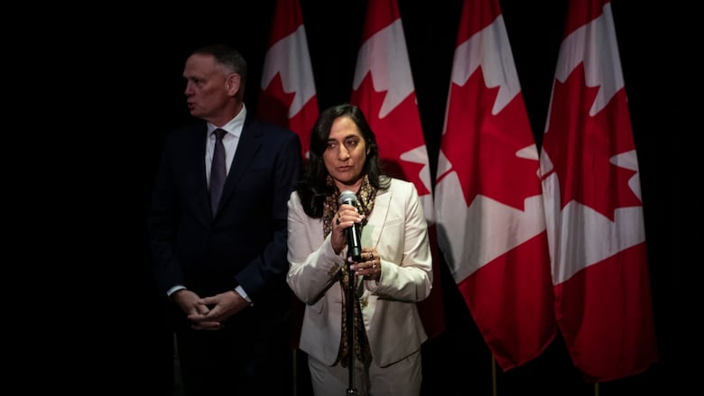 A woman speaks into a microphone as a man in a suit stands beside her. Four Canadian flags are drapped in the background.