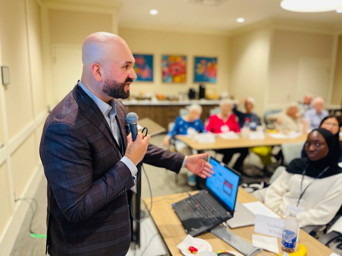 At the front of an indoor room, a man in a dark plaid suit and trim beard is seen speaking into a microphone as a mix of seniors and young adults listen in.