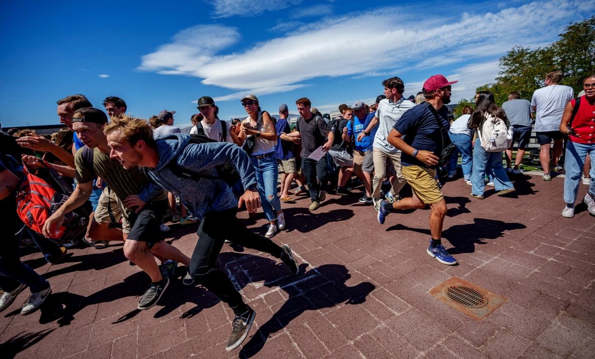 A crowd of people run away. Many look scared or nervous, and some are looking behind them at a point in the distance in the middle of the image. 