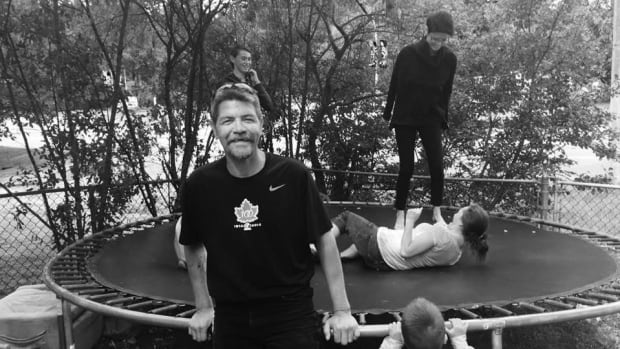 A man looks towards the camera, smiling, as children jump on a trampoline