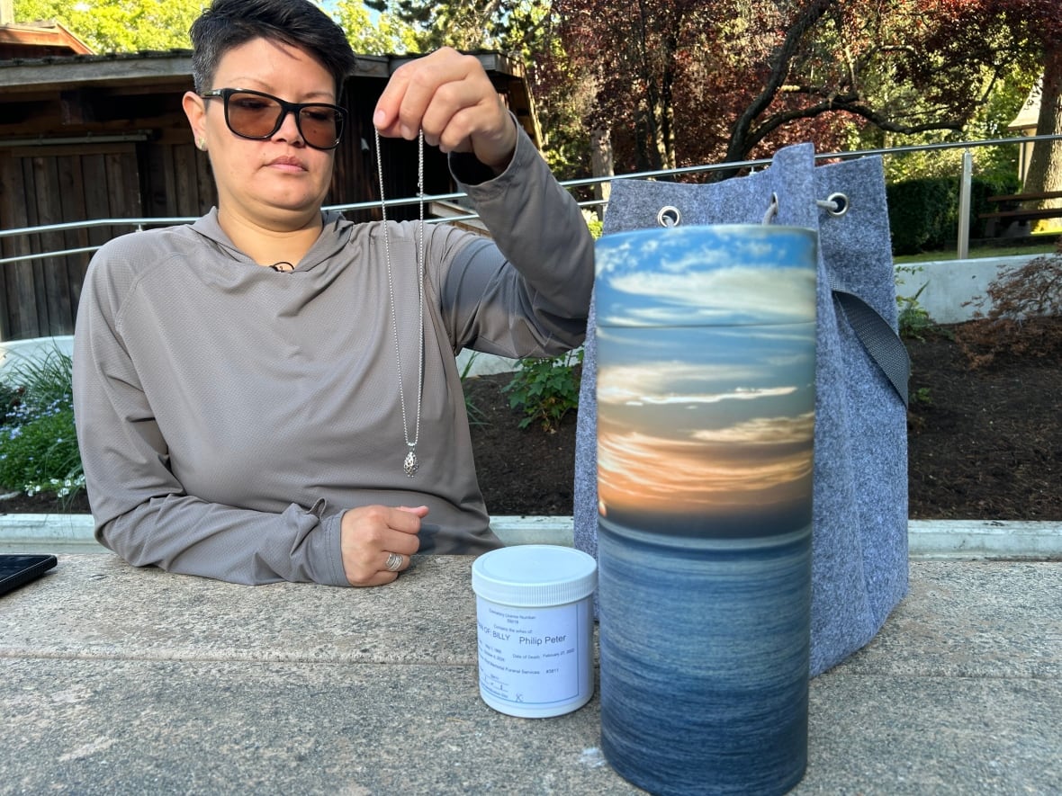 A woman holds up a necklace, next to two containers for ashes.