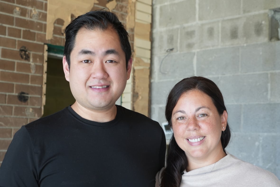 A man and a woman stand in a room stripped down to the cinder block walls