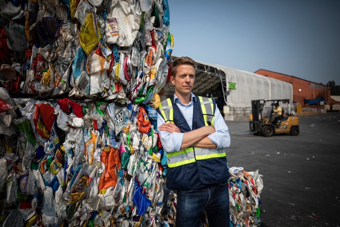 A tall man with blonde hair stands beside crushed and stacked plastic containers wearing a high viz vest.