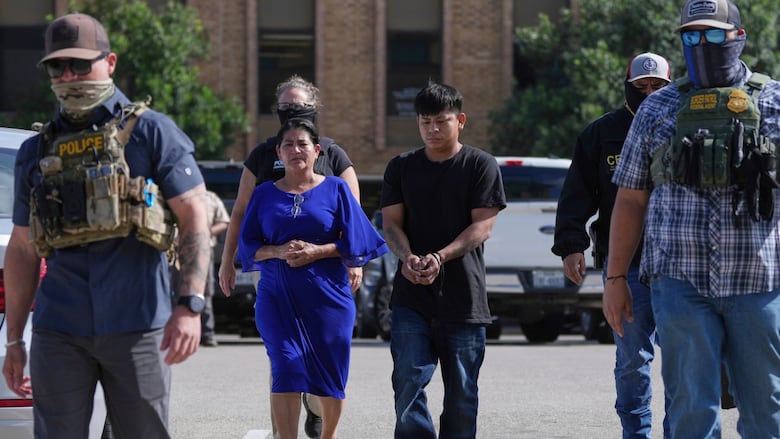A man and a woman, both handcuffed, walk between four masked federal agents.