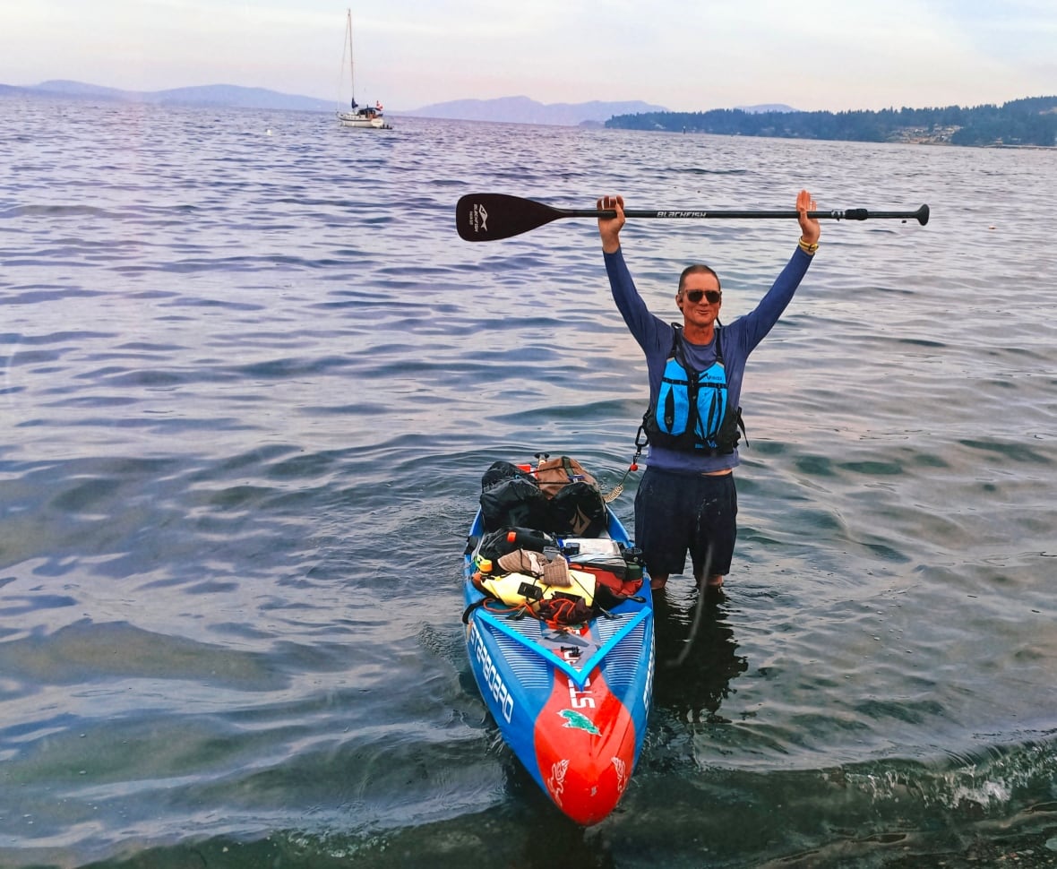 A person stands in the water next to a paddle board holding a paddle in the air.