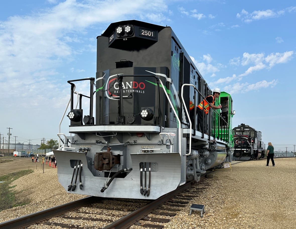 A large black and green painted locomotive sits on railway tracks. A man in an orange vest leans over the railing of the locomotive. 
