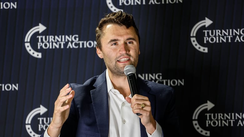 A man with dark hair wearing a navy jacket over a white collared shirt speaks into a microphone in front of a wall branded with a logo for Turning Point Action.