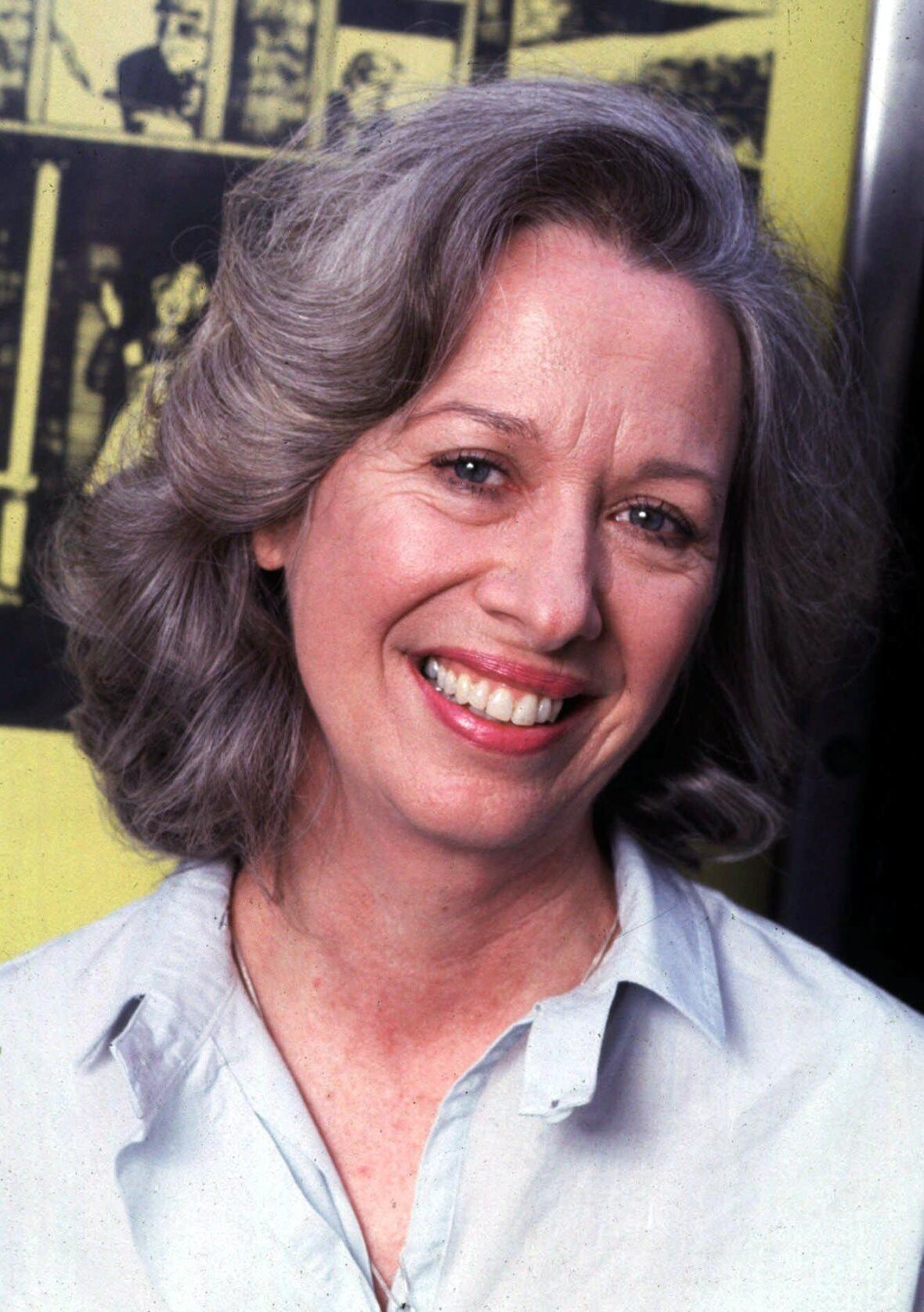 A woman with greyish hair and red lipstick is shown smiling while wearing a collared shirt in a closeup photo.