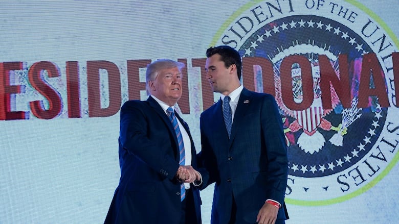 A young darkhaired man in a suit and tie shakes hands with an older man dressed similarly. Both are clean shaven. 