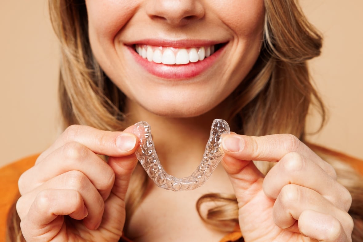 Nahaufnahme einer jungen Frau mit kurzem Haar, die ein orangefarbenes Hemd und legere Kleidung trägt und unsichtbare transparente Aligner in der Hand hält, ein Invisalign-Armschienenlächeln isoliert auf schlichtem pastellfarbenem hellbeigem Hintergrund.