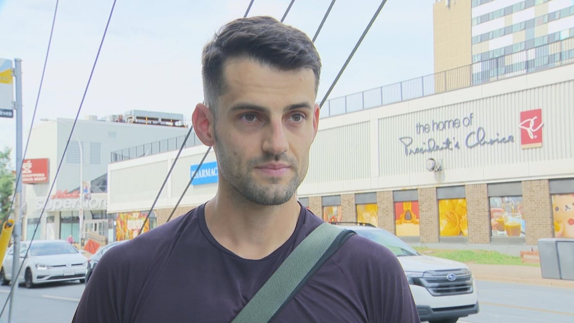 A person with short hair and a a short beard stands on a sidewalk by a road wearing a cross-body bag.