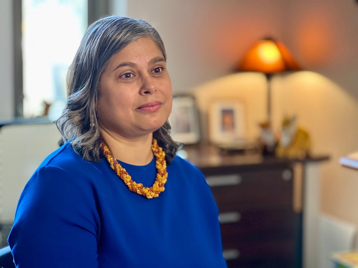 Angela Fernandez is seated wearing a blue shirt with a yellow and orange necklace.