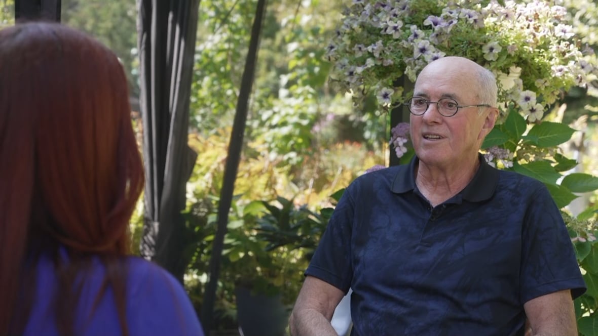 An older man in a blue shirt wearing glasses sits in a garden across from someone with long red hair.