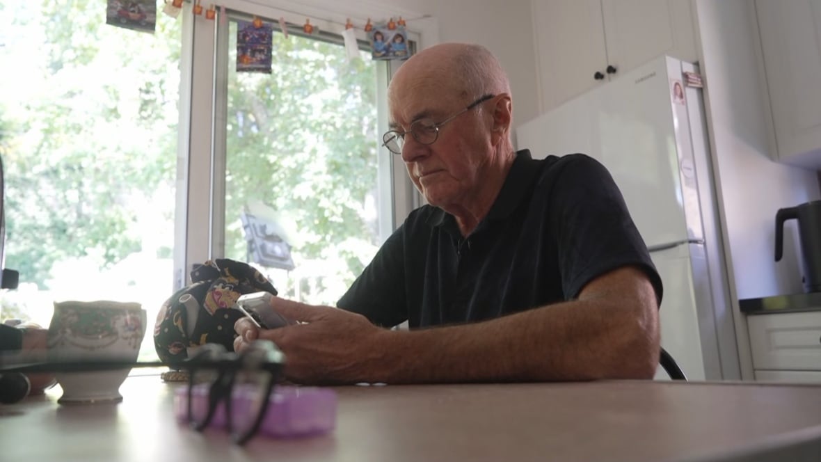 An older man wearing glasses and a blue shirt looks at his phone while sitting at a kitchen table.