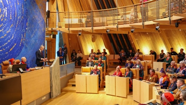 A wide view of a large room where people sit behind wooden desks facing a man at a podium.