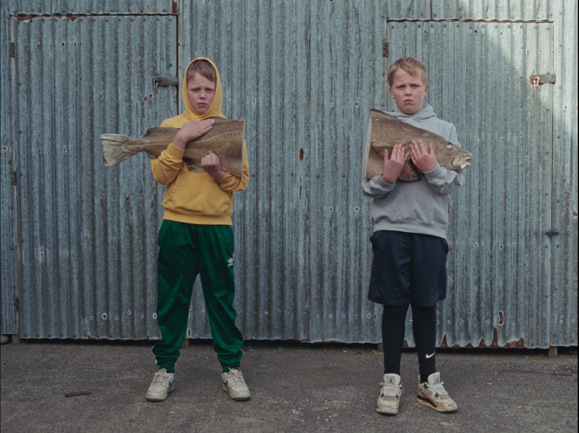 Twin boys stand slightly separated from one another. They hold two halves of a large fish split down the middle. 