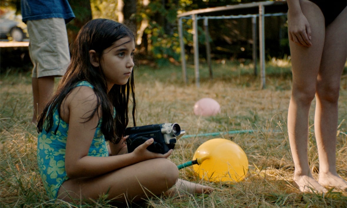 A young girl in a bathing suit holds a video camera while sitting on the grass.