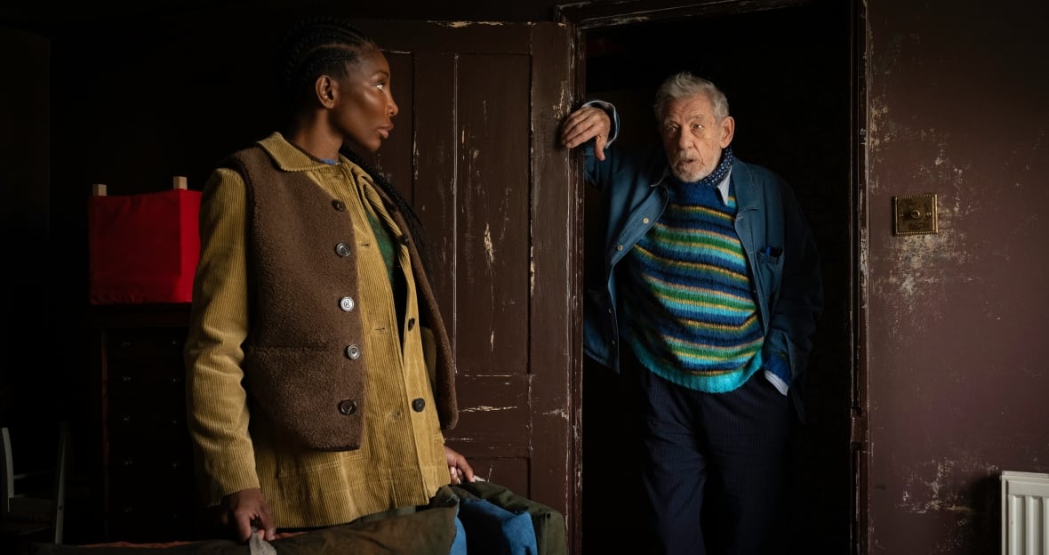 A woman stands looking questioningly at an older man leaning against a door frame. 