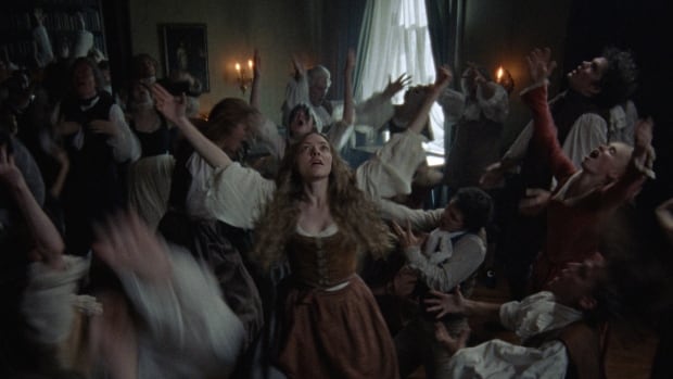 An ecstatic looking woman in period clothes dances in a large group. 