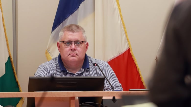 A man sitting behind a table with the Acadian flag in the background.