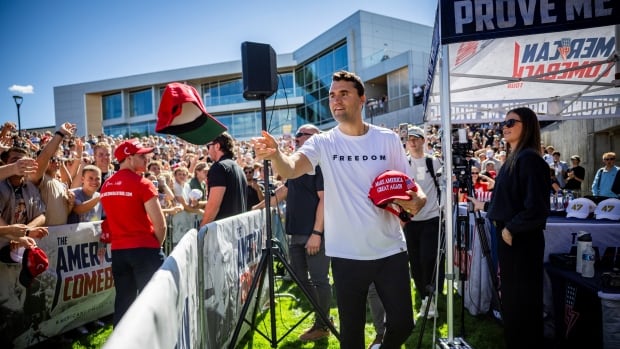 A man with dark hair wears a white t-shirt that says freedom as he tosses a red Make America Great Again hat to a crowd of people.
