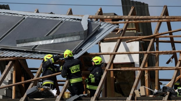 Firefighters in yellow helmets and uniforms examine the wooden frame of a house that has been damaged.