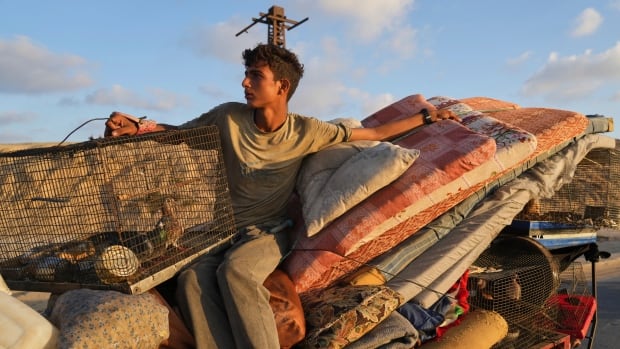 A young man holds onto a bird cage and other belongings atop a run-down vehicle.