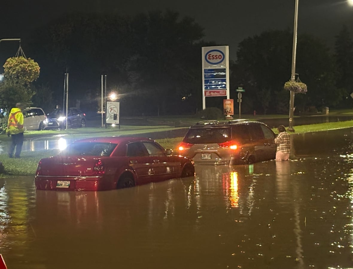 Vehicles are stranded in a flooded street, with water nearly to the top of the wheel wells.