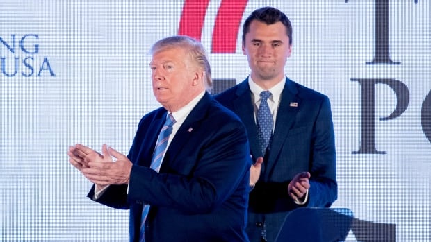 Two cleanshaven men in suit and tie clap while on stage. One is younger with brown hair, while the person in the foreground is a senior citizen, with blonde hair.