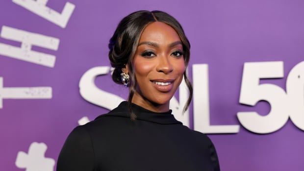A woman with dark hair thats pinned in an updo smiles for the camera in front of a purple background with white lettering