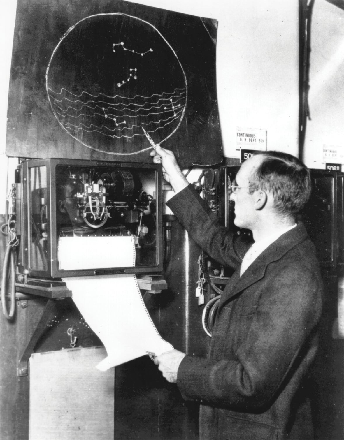 Une photo en noir et blanc d'un homme portant des lunettes à côté d'une sorte d'instrument d'où sort du papier.