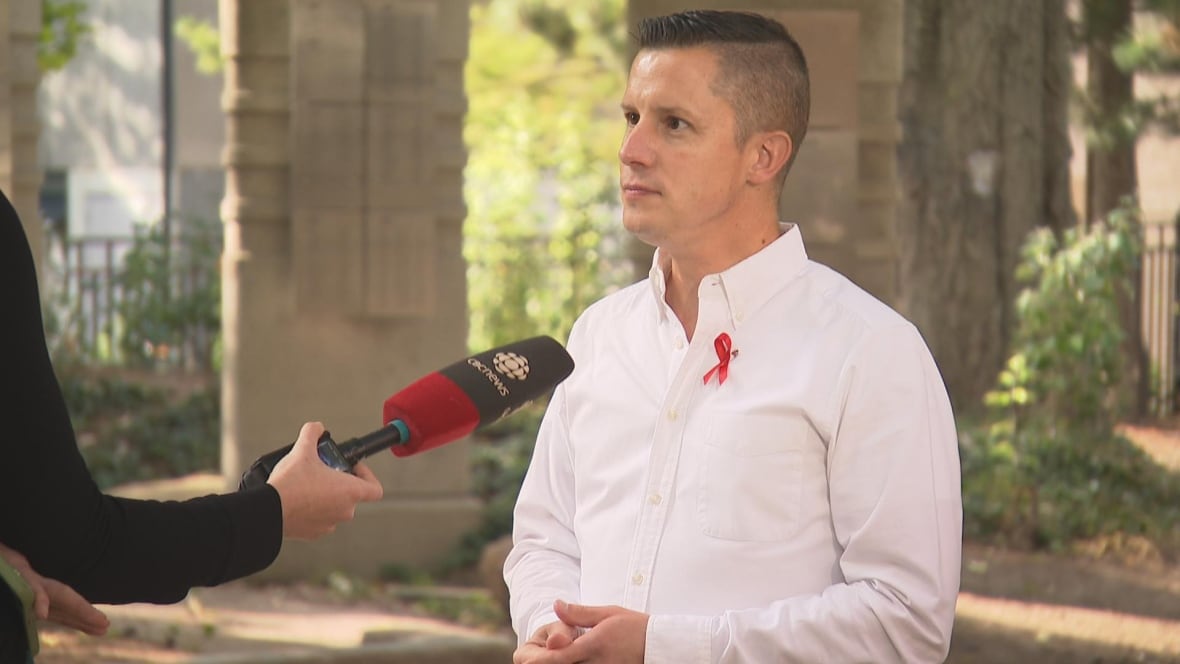 A man is wearing a white shirt and a red ribbon pin while talking to a reporter.