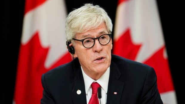 A man wearing a suit, red tie and glasses speaks. 
