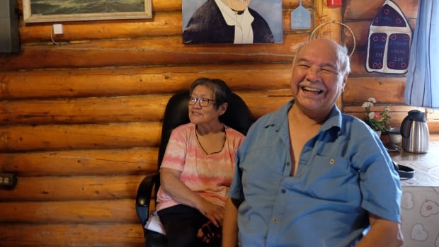 A smiling couple relaxes in front of a wall of a log house.