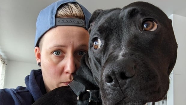 A blonde woman wearing a baseball cap put her arm around a large black dog. 