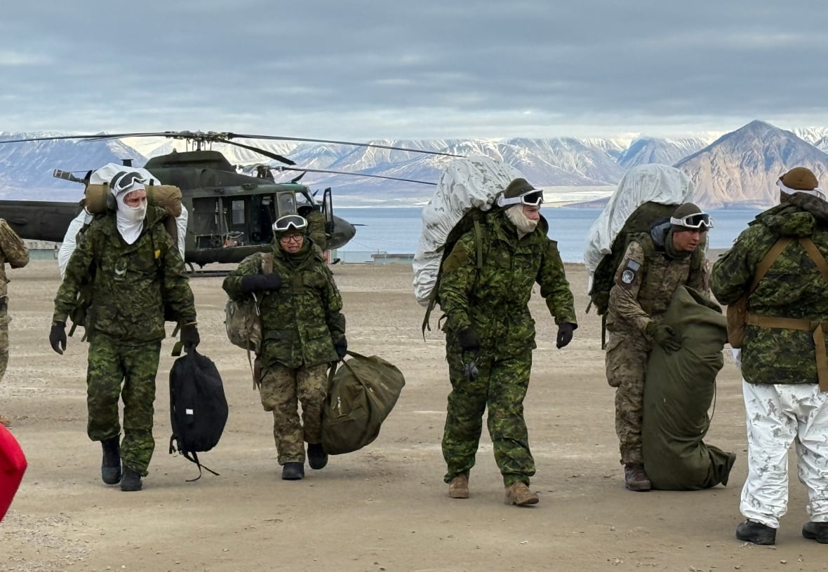 Five soldiers stand in arid landscapes in heavy winter clothes. There is a helicopter and some mountains in the background.