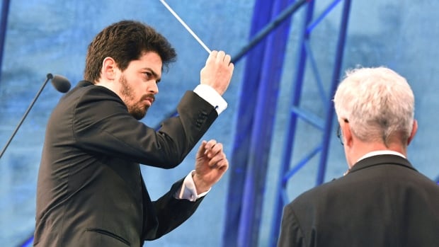 a serious looking man in a suit waves a conducting baton, conducting an orchestra that is not pictured