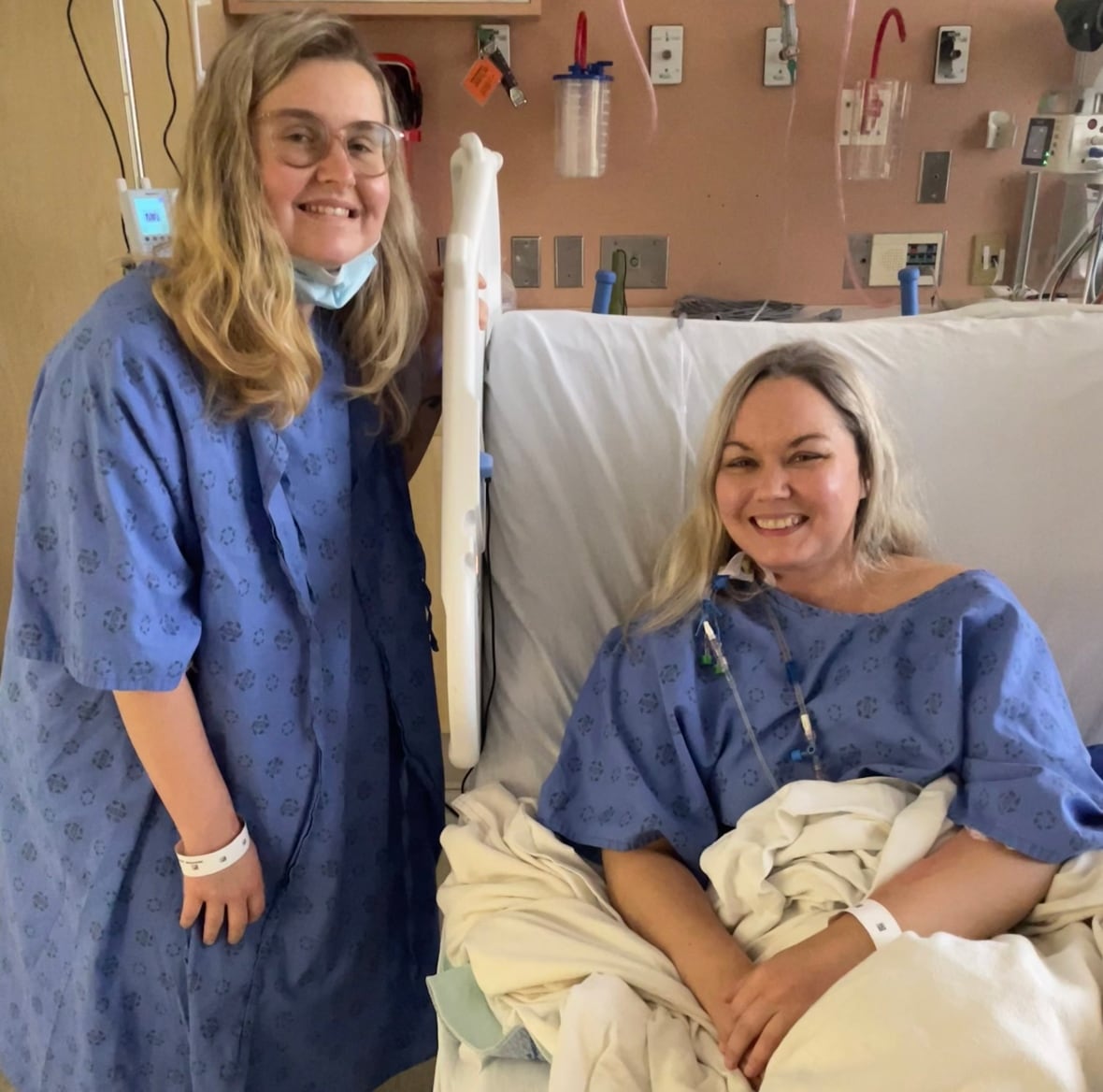 A smiling woman wearing a medical gown, standing beside another smiling woman wearing a medical gown, lying in a hospital bed.