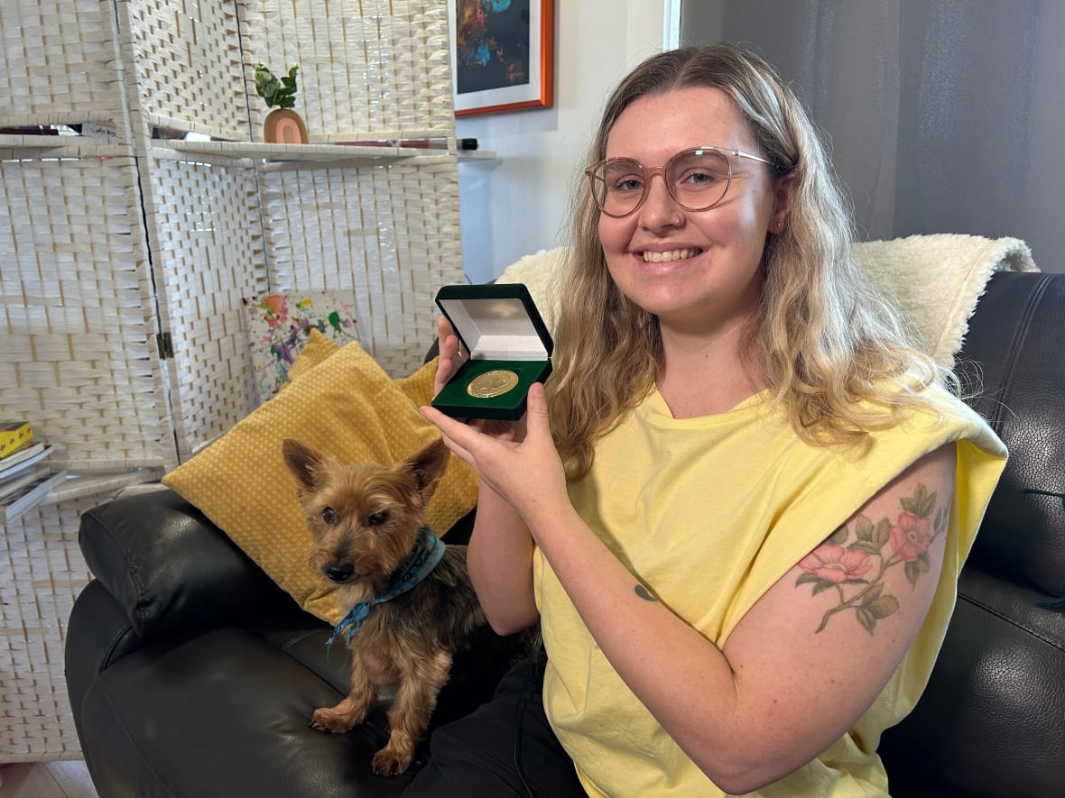 A smiling woman, sitting on a couch beside a small dog, and holding up a small open box that contains a gold medal.