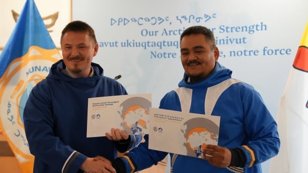 Two men stand holding up papers and shaking hands.