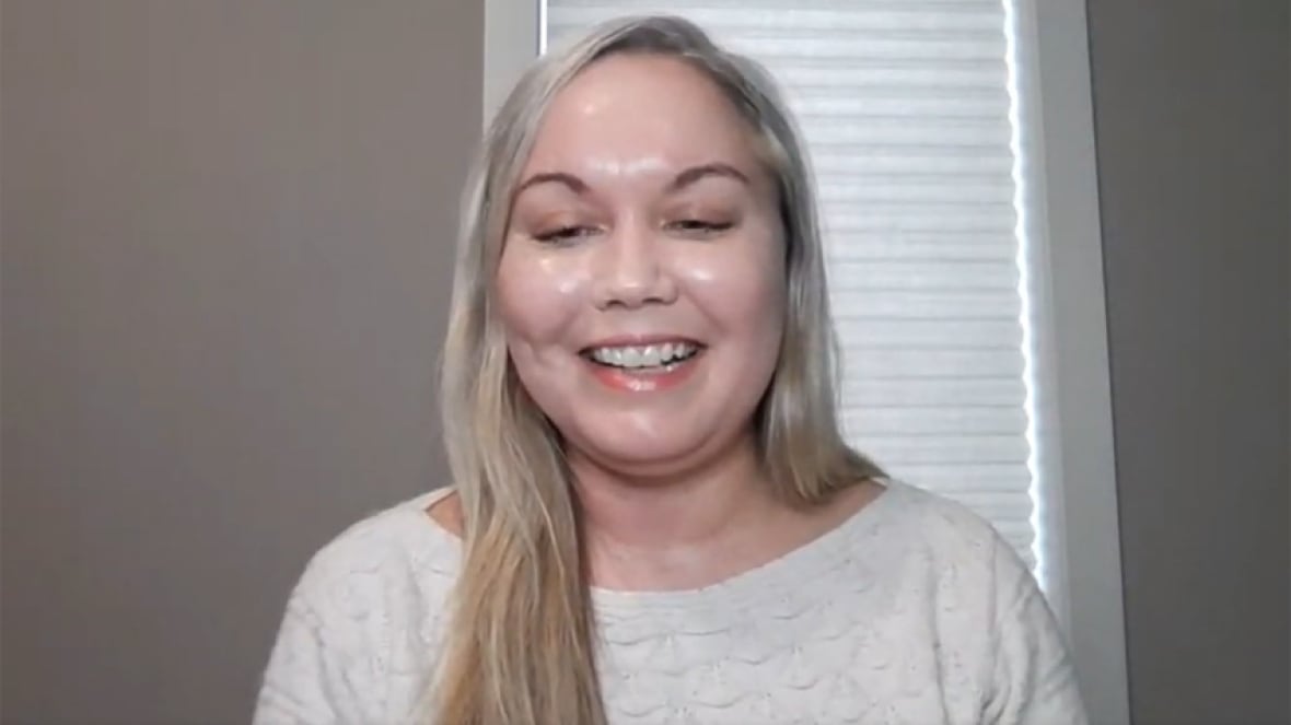 A smiling woman with long, straight blond hair, wearing a white sweater, sitting in front of a window with a white blind pulled down.
