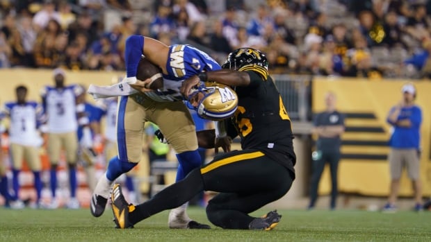 A player in black wrestles a ball carrier in blue, gold and white to the ground.