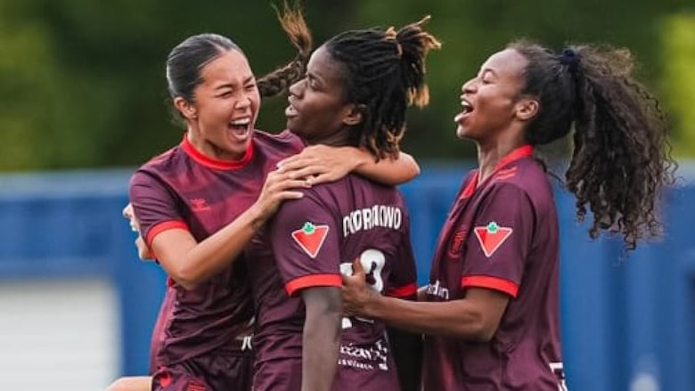Two female soccer players smile while wrapping their arms around a teammate in celebration on the field.