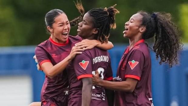 Two female soccer players smile while wrapping their arms around a teammate in celebration on the field.